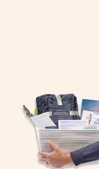 Hand holding a white woven Pearl Project gift set basket filled with a navy chenille blanket, "The Unfolding" grief and loss guidebook, question cards, artwork, tea, and eucalyptus.