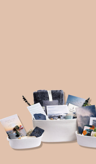 Pearl Project gift sets in a white woven basket, holding a navy throw blanket, the "Facing the Storm" therapist-written guidebook, question cards, framed nature artwork, a candle, and greenery.