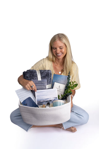 A smiling woman holds a white woven Pearl Project gift set on her lap, containing a navy blanket, "The Unfolding" guidebook, a watercolor seascape print, a candle, and eucalyptus.
