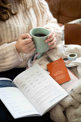 Woman holding coffee mug on couch with The Pearl Project mental health kit including workbook and question cards