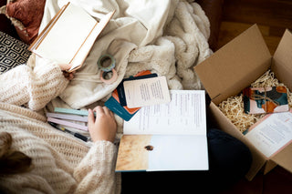 Overhead view of a person wrapped in a knit sweater and blanket, using a Pearl Project gift set — an open guidebook, "Into the Storm" reflection question cards, and a journal — with the open kraft gift box and thank-you card nearby.